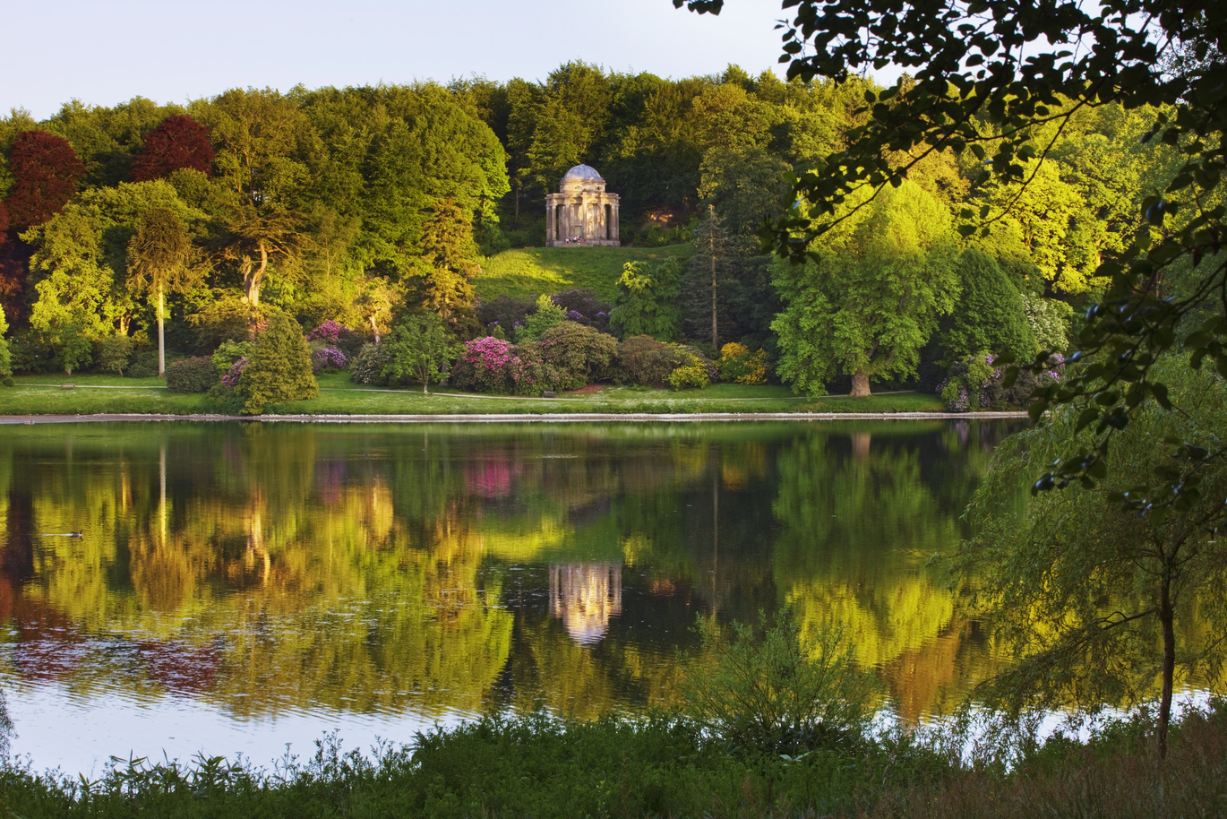 The 18th century Temple of Apollo reflected in the lake surrounded by many green trees with the sunlight shining down.