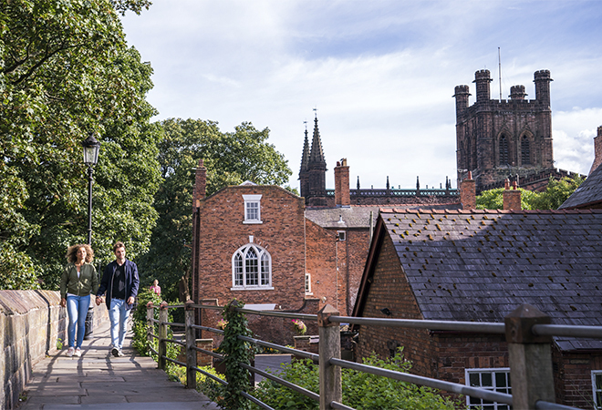 "A male and femme walking along a pavement with a wall to one side and a low rail on the other with Chester City Centre in the background"