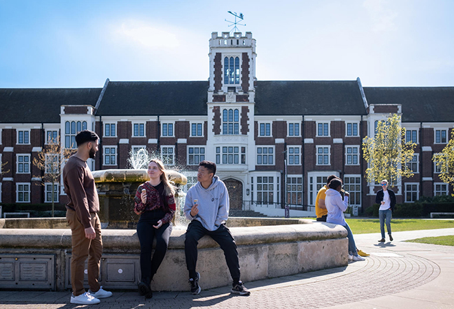"Two groups of students socialising next to a fountain outside the front of Hazlerigg Hall. It is a large brick built building with a tiled pitched roof and white borders around its many windows."