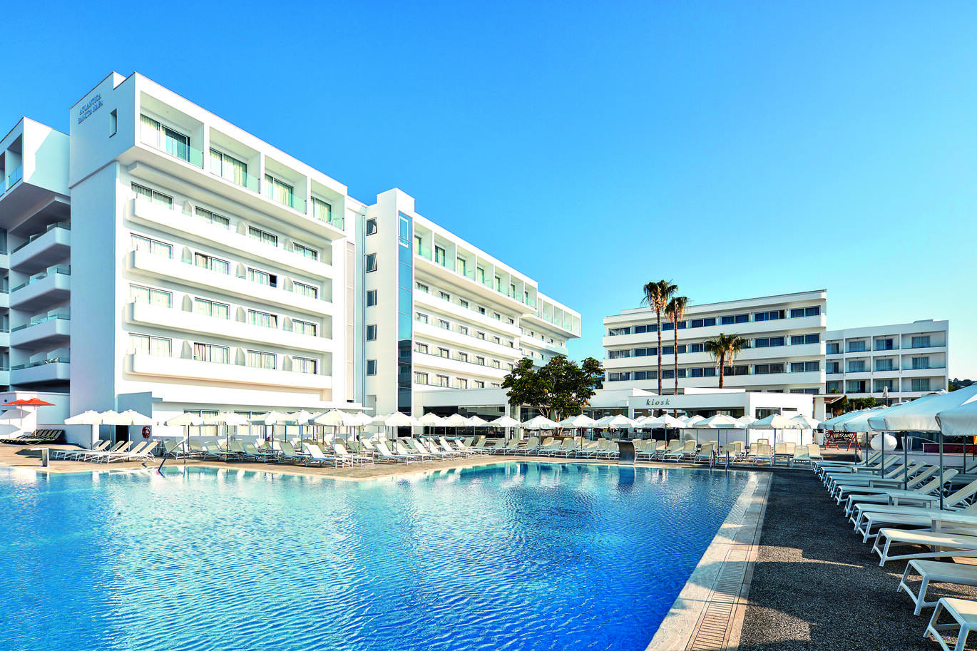 View of the pool area surrounded by white sun loungers and umbrellas, with the tall white hotel buildings behind and the clear blue sky above.