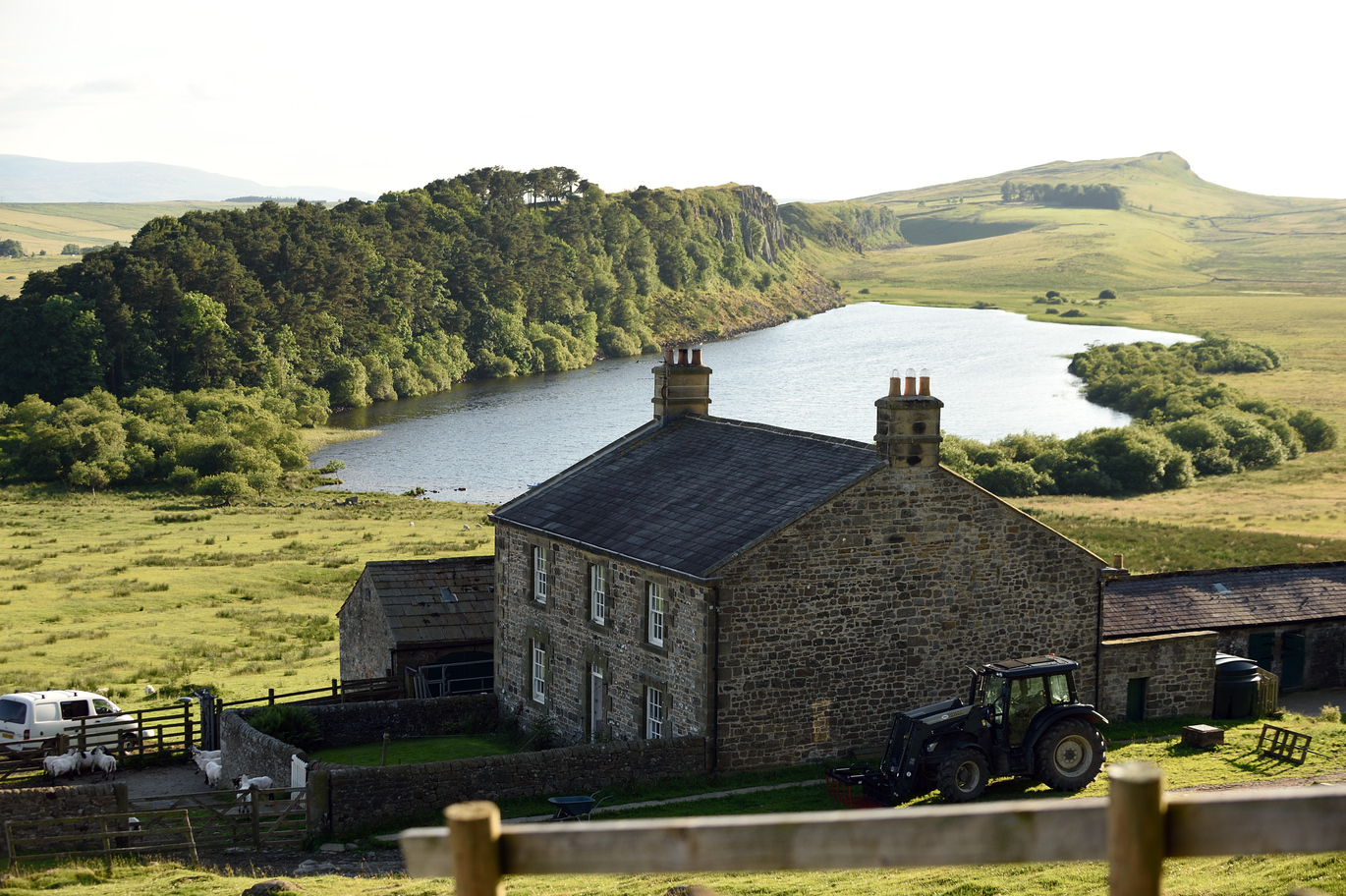 View of Hadrian's Wall and Housesteads Fort with the lake and fields behind the house and a tractor parked next to the house.
