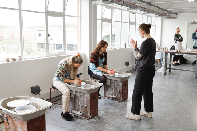 'two people sat at pottery wheels while being taught by a third person' 