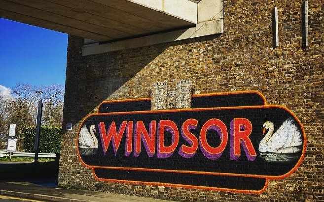 "Photo of a painted sign on a brick wall saying Windsor in red writing with a purple edge to the writing, swans painted in white either side of the word, a black background and Windsor Castle above the word"