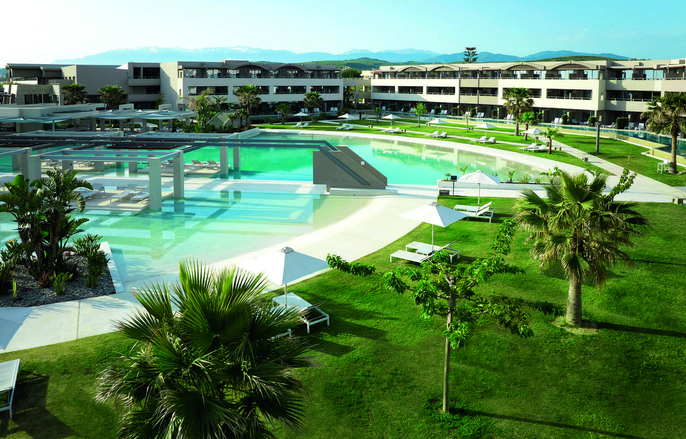 View of the pool area surrounded by lush green lawns, palm trees and sun loungers with the hotel building surrounding behind, and mountains just visible in the background.