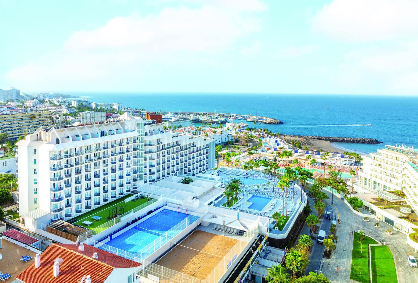Aerial view of the hotel complex with pools, a tennis court and a volley ball court in front of the large white hotel building. The beach and sea are visible over the other side of the road 