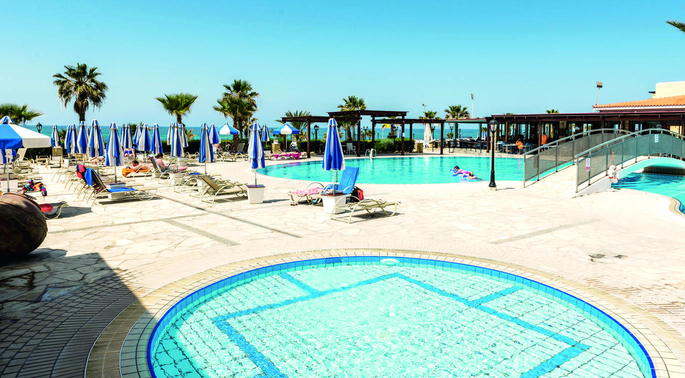 View of the pool area with one big main pool with a bridge over and one small round pool next to it, surrounded by deck chairs and umbrellas, with palm trees behind and the sea visible in the distance on a clear, sunny day.
