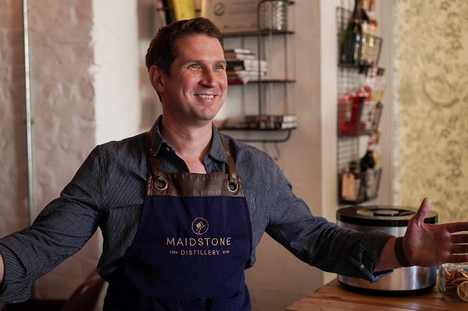 "Photo of a man standing with his arms out to each side, smiling and wearing an apron with the words Maidston Distillery on. There are bottles in the shelf in the background."
