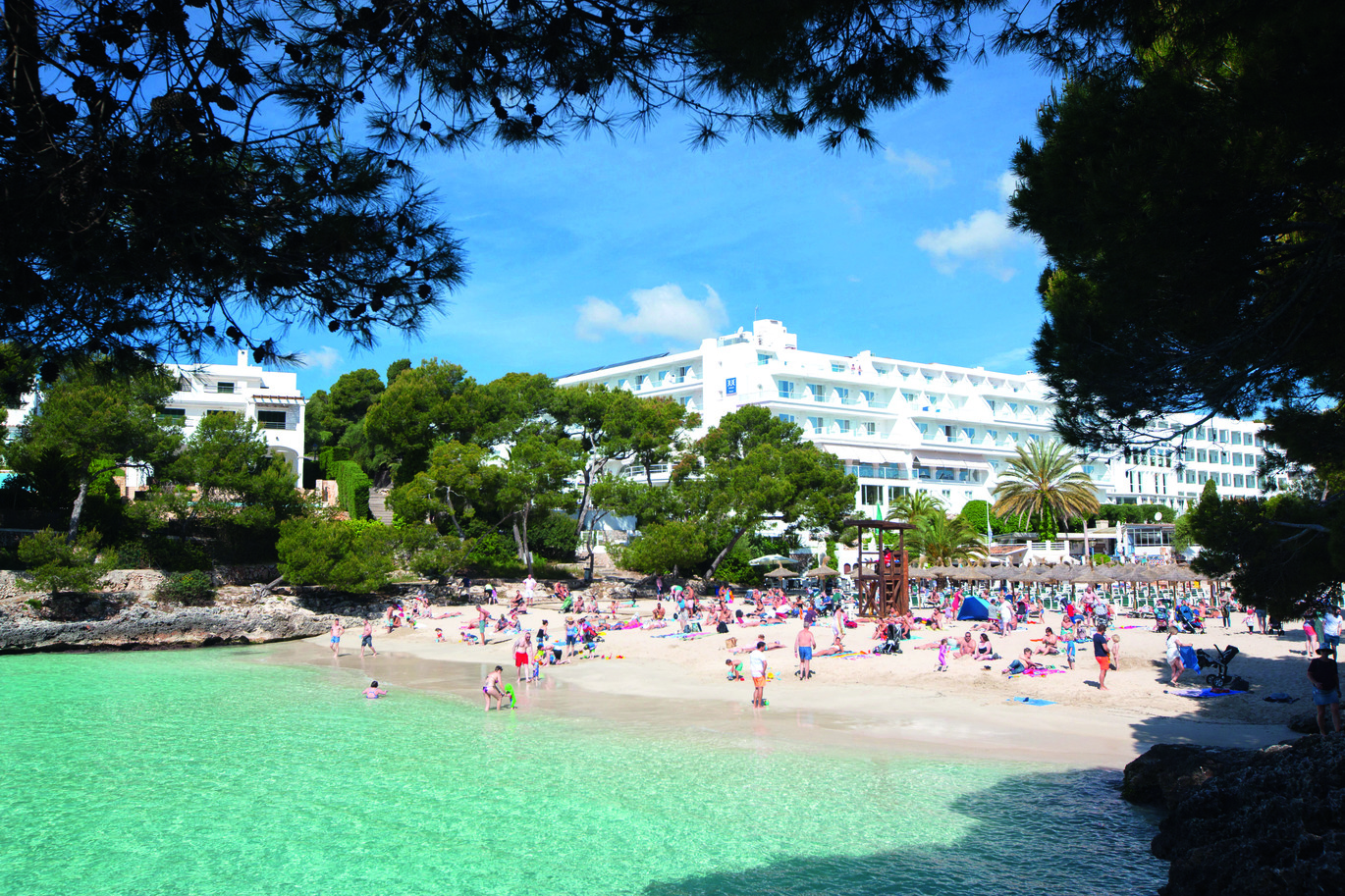 View of the beach area with the beautiful blue sea and many people around, surrounded with trees and the large, white hotel building behind.