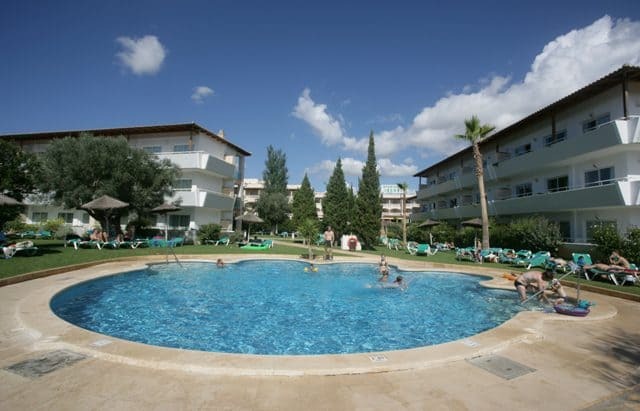 View of the hotel building with the pool in front with people in it, surrounded by green lawn with seating also with people around.