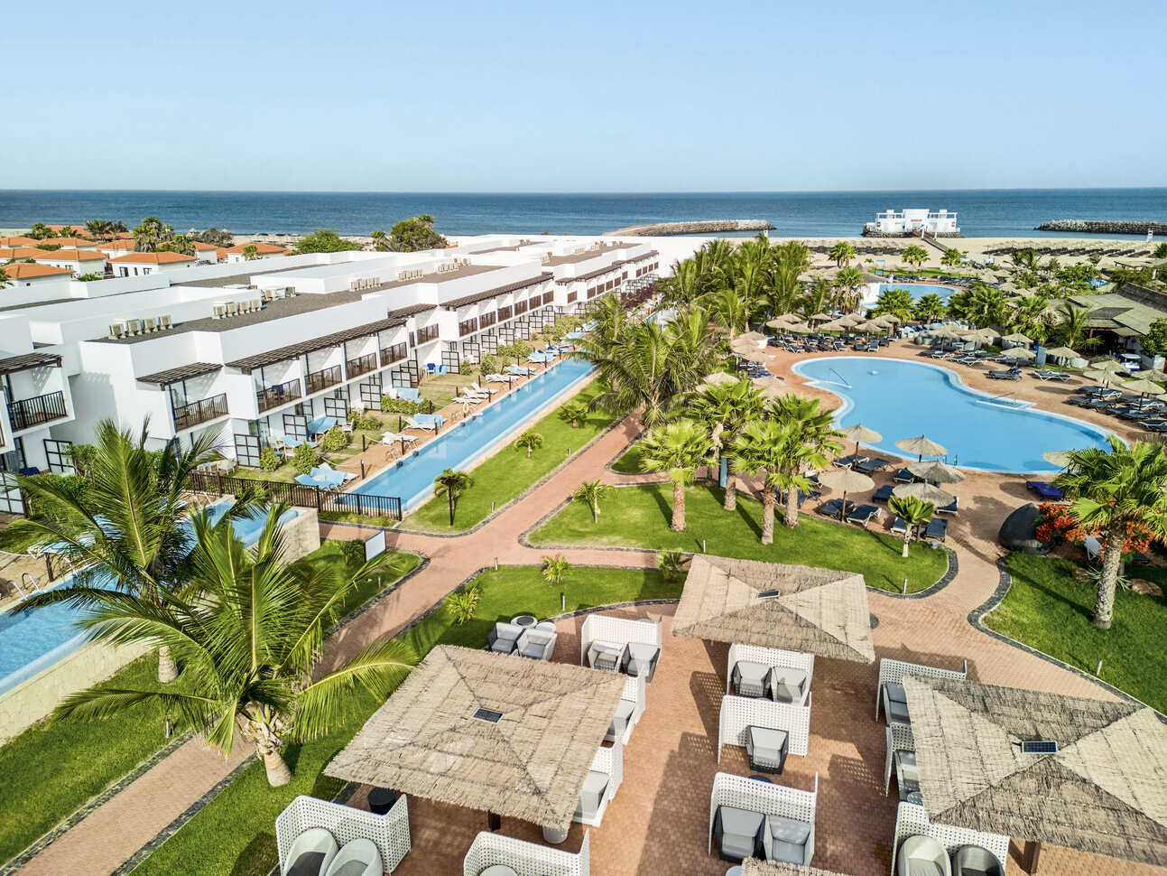 Aerial view of the hotel complex with the main pool and swim up rooms visible, with a seating area at the front and lawns and palm trees all around. The sea is visible in the background with the blue sky above.