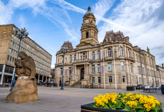 The front of the town hall with stairs leading up to the entrance. A very large stone building with many windows and a clock at the top. A large brick building is to the left of the town hall made up of mostly windows. In front of the town hall is a market square with a sculpture of a man holding up a woman. There is a pot of yellow flowers in the bottom right of the image and the blue sky with clouds in above.