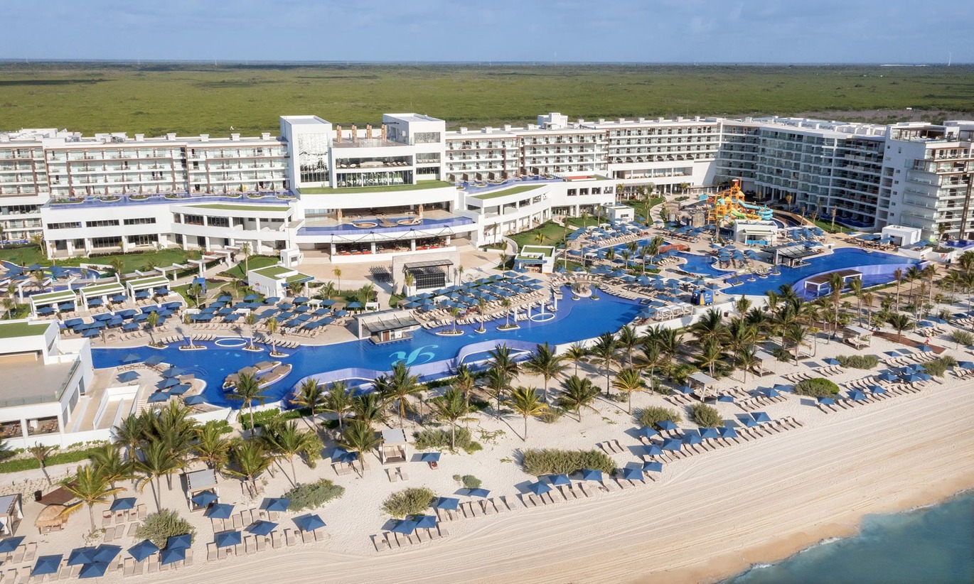 Aerial view of the hotel complex right on the beach with palm trees separating them. With 2 large swimming pools and a play area with water slides in one. the pools are surrounded by many sun loungers with blue umbrellas and the large hotel building behind with empty fields directly behind.