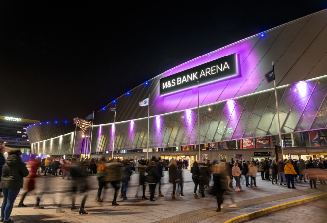 "Lots of people gathered outside entrance to a building with the sign lit up with a purple back light saying M&S Bank Arena"