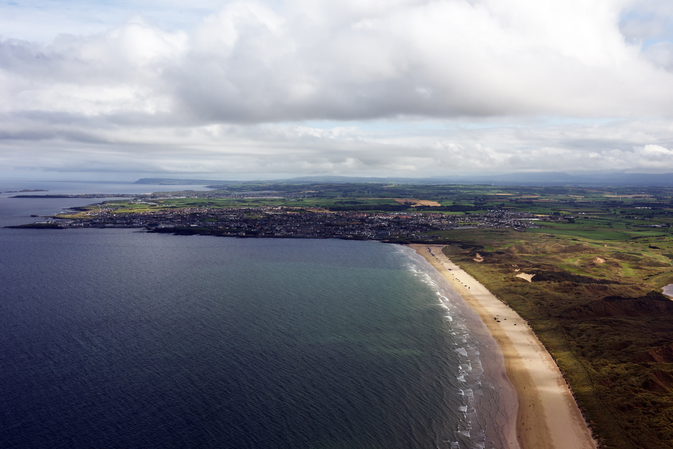 Aerial view of the beach and sea at Portstewart Strand with greenery and hills directly behind the beach and a village full of houses to the side right on the sea edge.