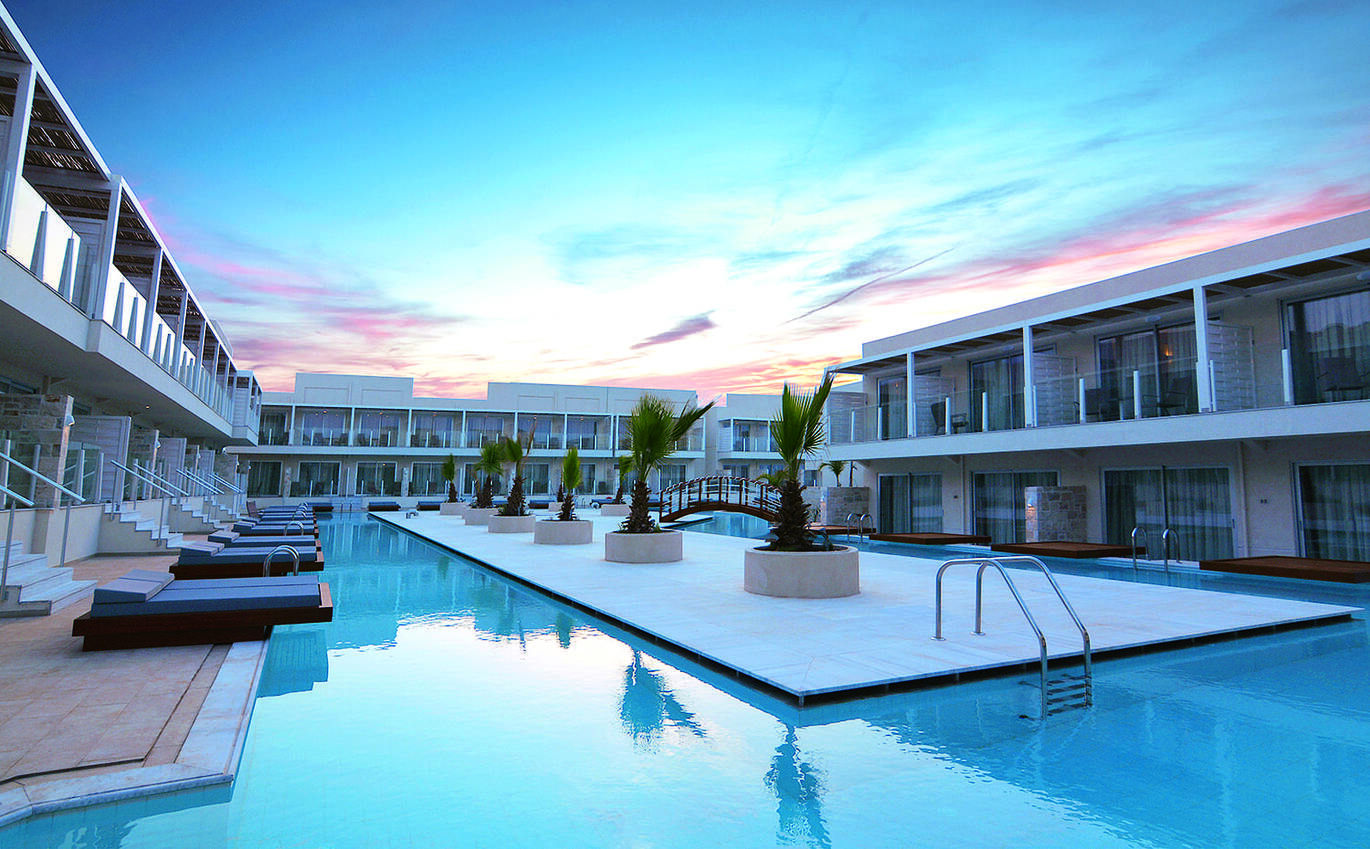 Sunset view of the pool area with an island in the middle and a bridge to the other side. With sun beds overhanging the pool and rooms directly behind.