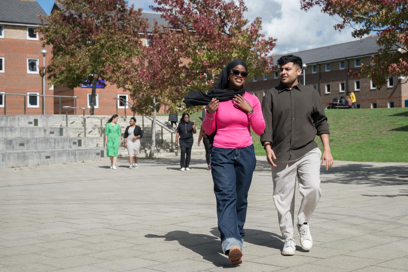 "Two people walk side by side across a paved campus courtyard, with other people in the background. Trees with autumn-coloured leaves and modern brick buildings line the open space on a bright, sunny day"