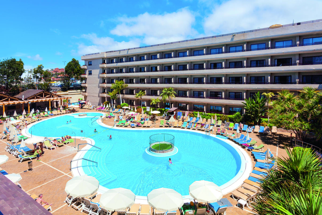 Aerial view of the pool area with many sun loungers and umbrellas around and an undercover hut with seating. With the hotel building surrounding.