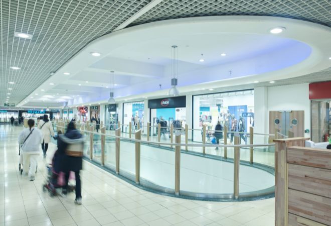 An indoor shopping mall with a modern design. The flooring is tiled, and the ceiling features a curved, grid-like pattern. There are various retail stores with glass storefronts on the right side, and a wooden railing on the left side separates the walkway from another level of the mall.
