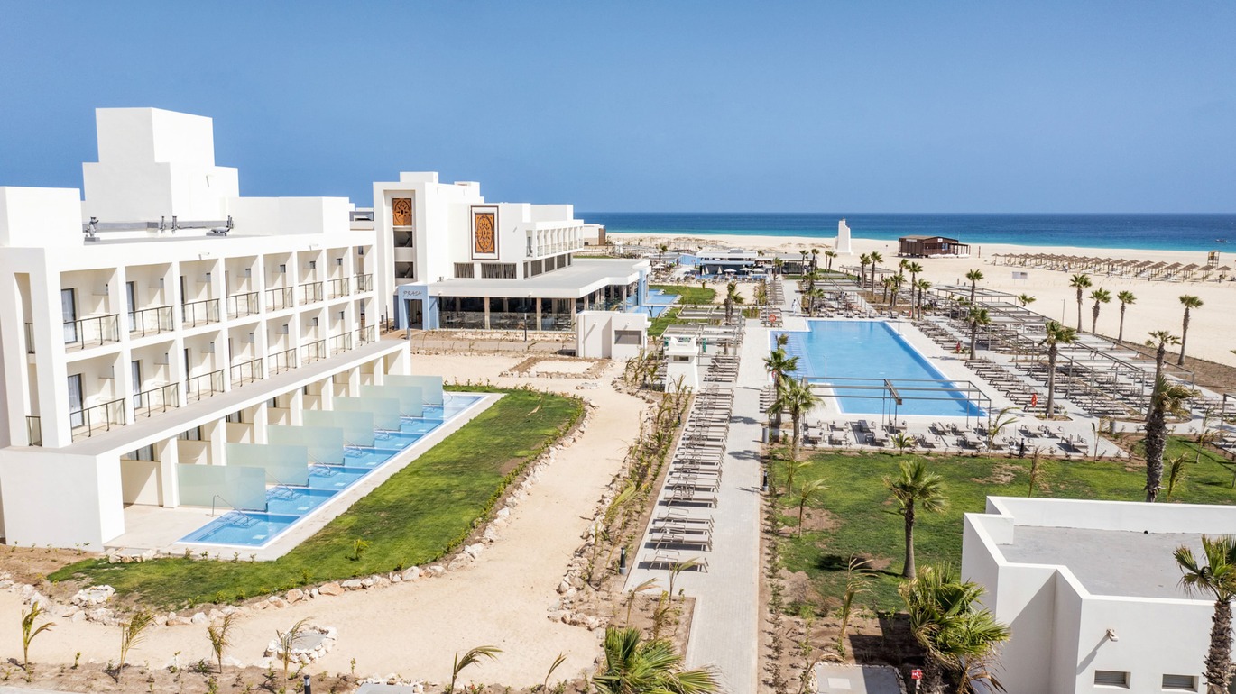 Slight aerial view of part of the hotel with rooms with private pools and the main pool surrounded by many, many neatly lined up sun loungers and the white sand beach behind with the sea just in view on a lovely clear sunny day.