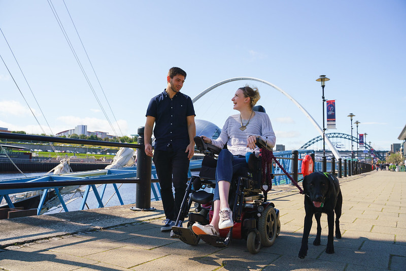 "Female electric powered wheelchair user and male walking a black Labrador along the Quayside"