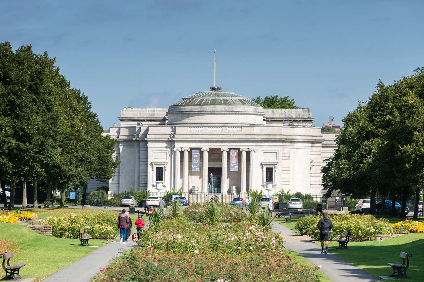 The image shows the Lady Lever Art Gallery; a historic building with a glass dome, surrounded by lush greenery and colourful flowerbeds, under a blue sky.