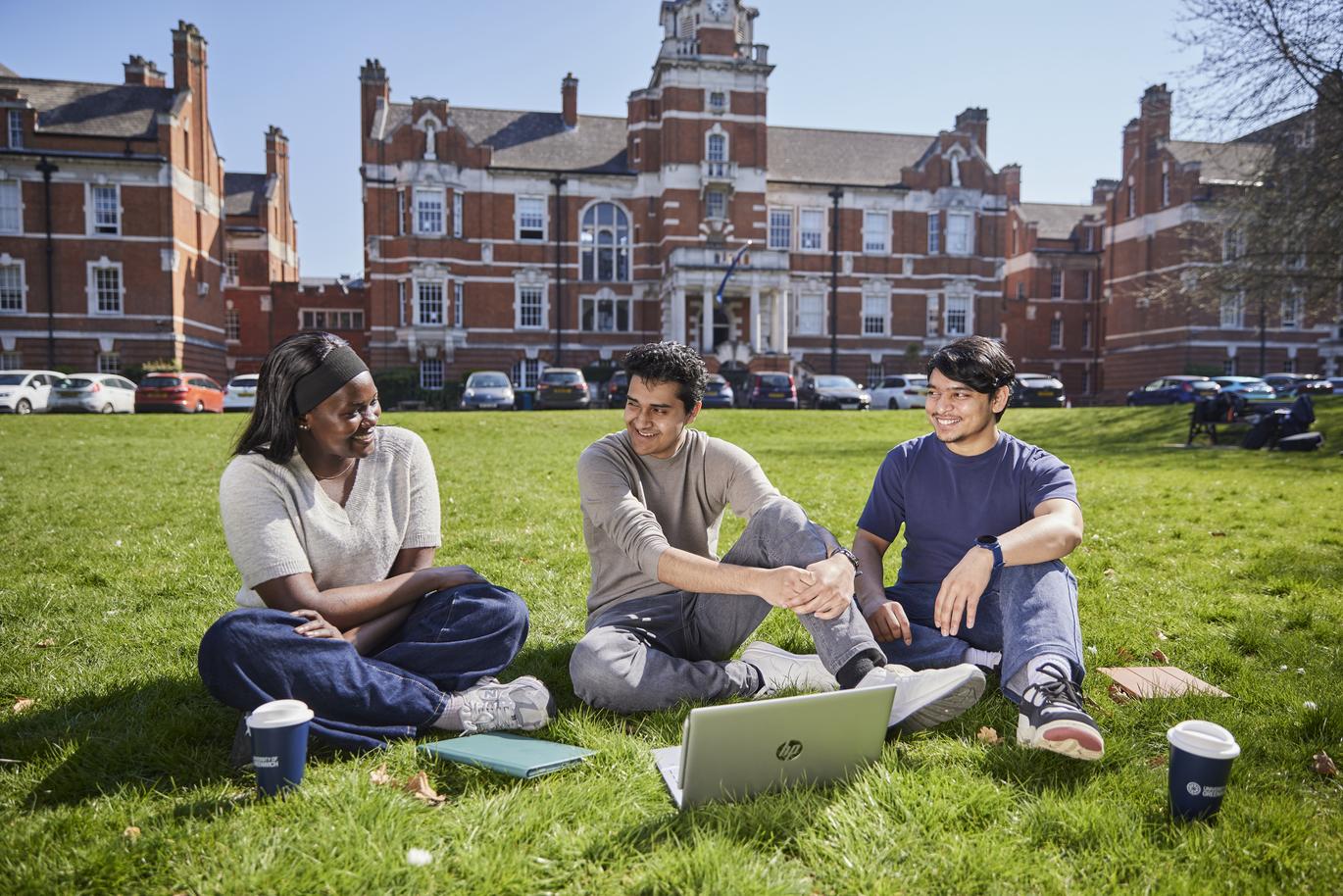 " Three people sit on a grassy lawn in front of a large red-brick university building, with a laptop and notebooks placed on the grass between them. The group appears to be studying together on a bright, sunny day"
