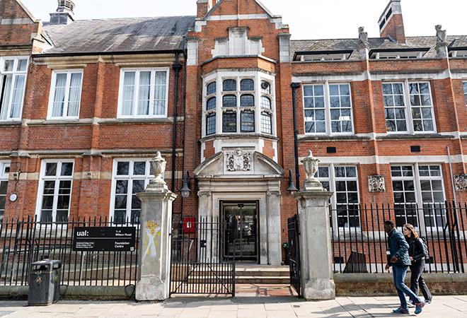 “Front view of Camberwell College of Arts Wilson Road campus, showing a historic red-brick building with large white-framed windows, a central stone entrance with decorative detailing, and black metal railings along the sidewalk. Two people are walking past the entrance.”