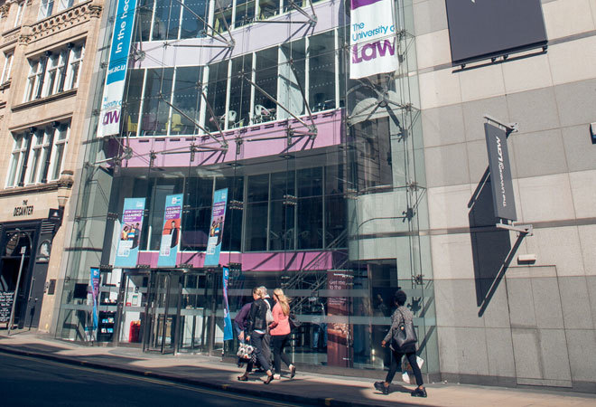 Photo taken from the road, of the university of law in leeds. The front of the building is all glass, and you can see inside each level. 4 pedestrians walk past.