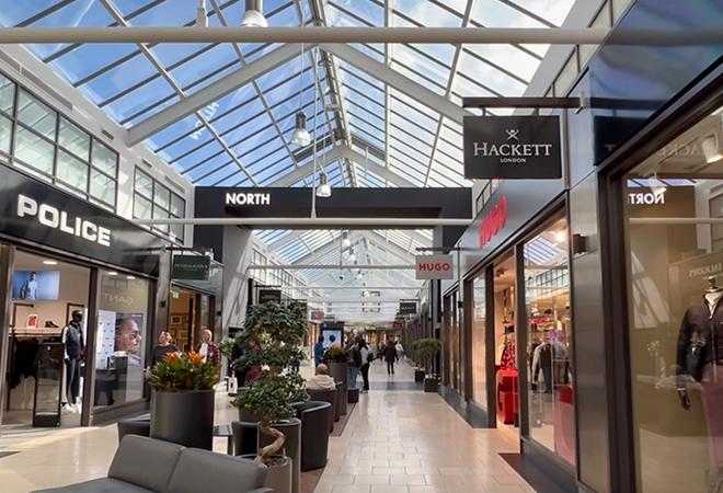 "Interior of a modern shopping mall with a glass ceiling, featuring stores like 'POLICE' on the left and 'Hackett London' on the right. A 'NORTH' directional sign hangs from the ceiling, with shoppers walking along a path lined with potted plants and seating areas."