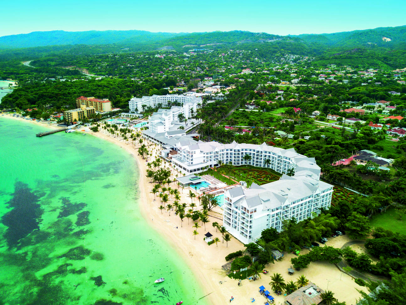 Aerial view of the large hotel complex with large white hotel buildings, swimming pools and lawns right on the beach front with the green sea, all surrounded by many trees, other buildings in the background and hills at the back.