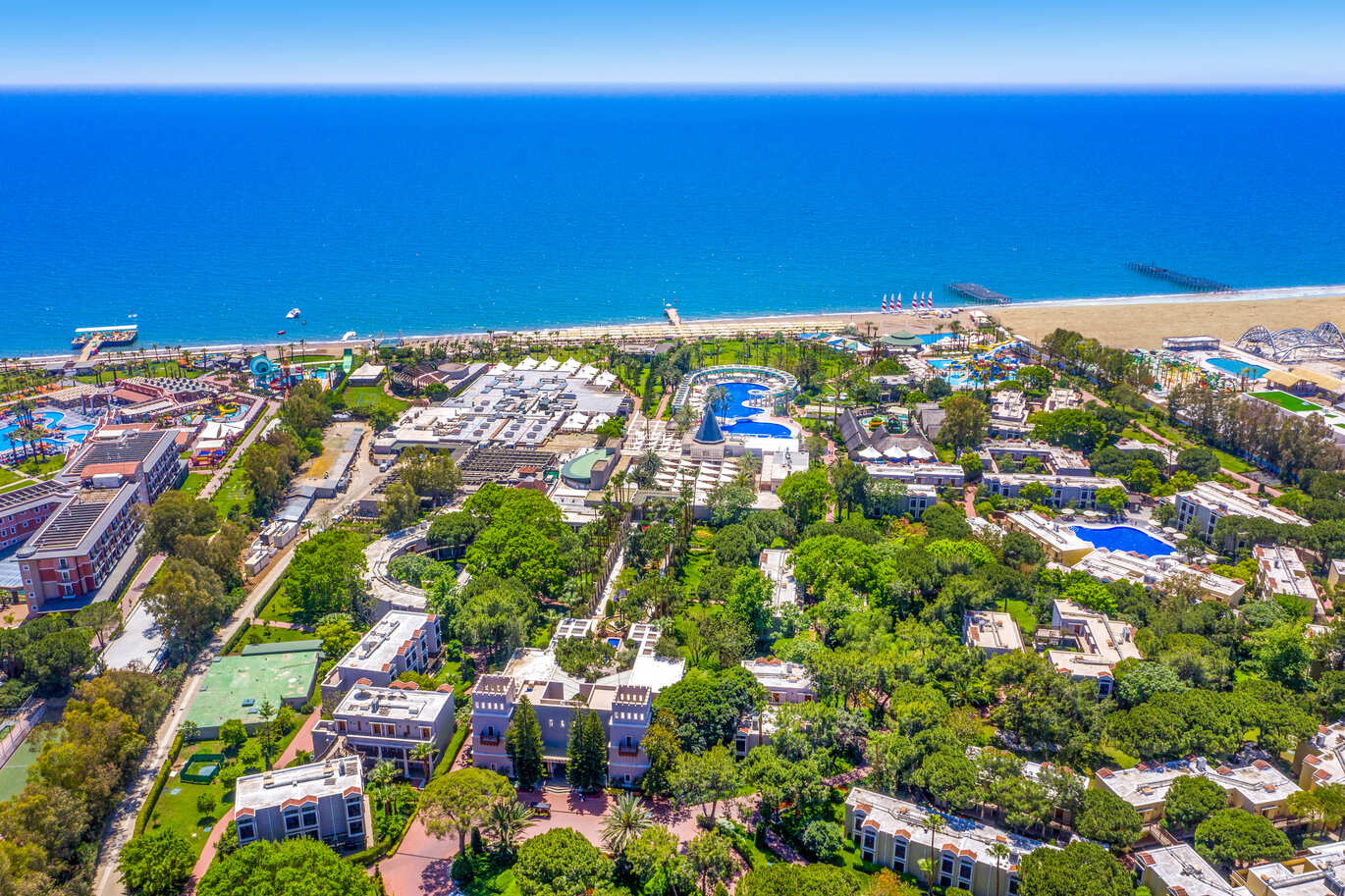 An aerial view of many different hotel complexes with blue pools surrounded by many trees with the beach in the background and the lovely blue sea on a bright, clear, sunny day.