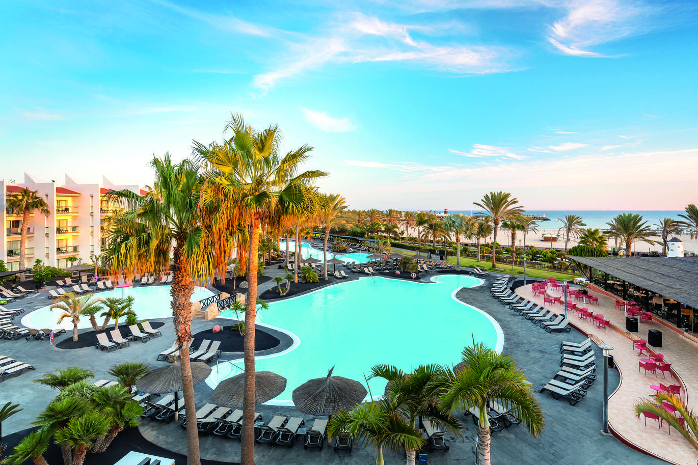 Aerial view of the pool area surrounded by many sun loungers, with lots of palm trees around and a small bridge over the pool. With a bar seating area to the right of the pool and the beach and sea in the background with the sunset reflecting on the hotel building.