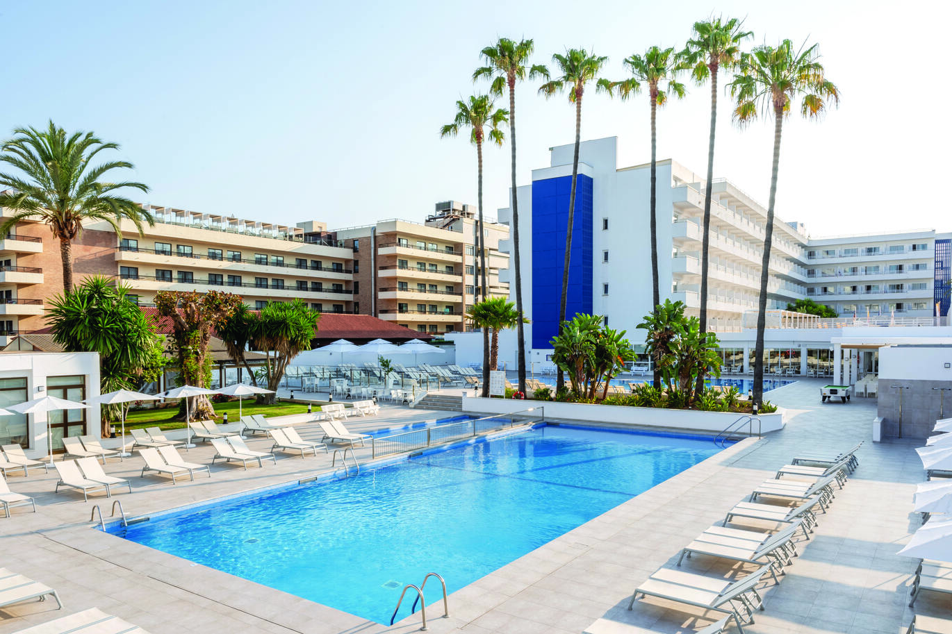 Hotel buildings showing a cluster of rooms with balconies with some palm trees in front. The pool in front with white sun loungers neatly lined up around the pool with white umbrella's above some of them. Lovely clear sky shown above.