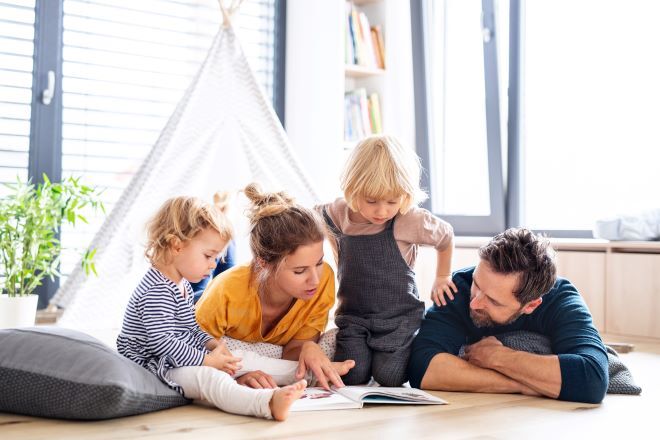 "A photo of a family, reading from a book together. A man and a woman are lying on their fronts, with their two children sitting next to them."