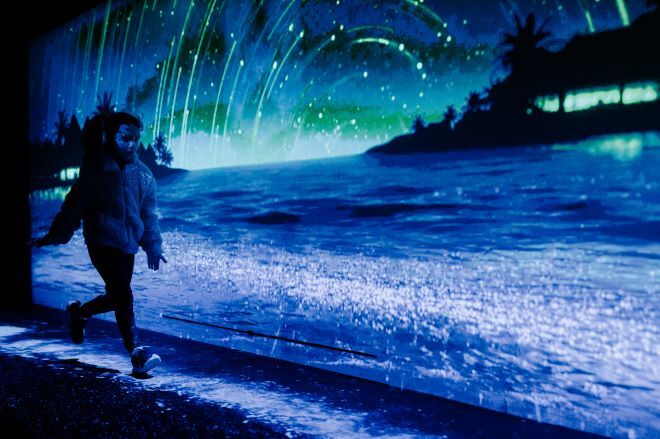 "A person in dark clothing walks in front of a large screen showing a digital beach scene at night, with waves, silhouetted palm trees, and streaks of light resembling shooting stars or auroras in the sky. The person's face is obscured by shadows."