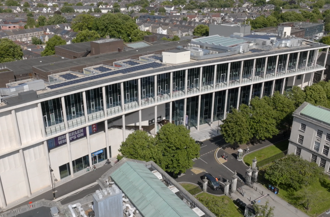 "Drone photo of a Cardiff University building, white building over three floors with a balcony walkway. Trees line the road in front."