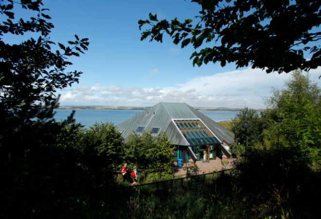 "Montrose Basin. Pyramid shaped silver building with bushes and trees in foreground. Mostly blue skies with some white clouds in background."