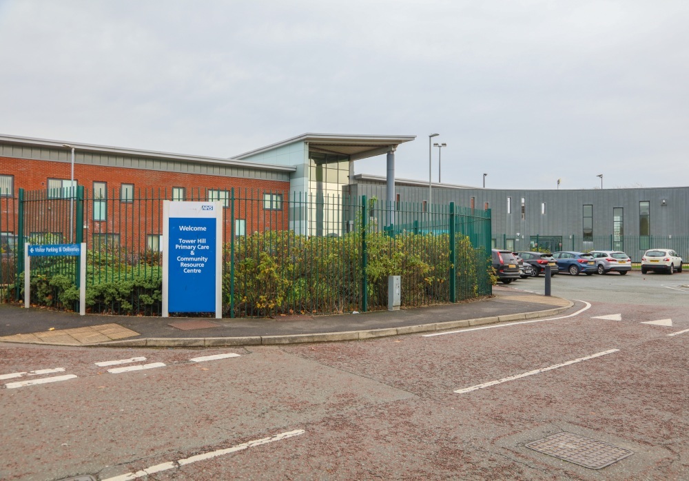 "A large two storey building - half brick built, half panelling with a car park and tall metal fence outside the front. There is an NHS sign which reads 'Welcome to Tower Hill Primary Care & Community Resource Centre."