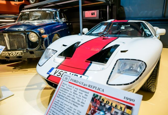 "Grampian transport museum. White care with red stripe up the middle of car. Plaque that reads Ford GT40 Replica. Vintage blue car in background."