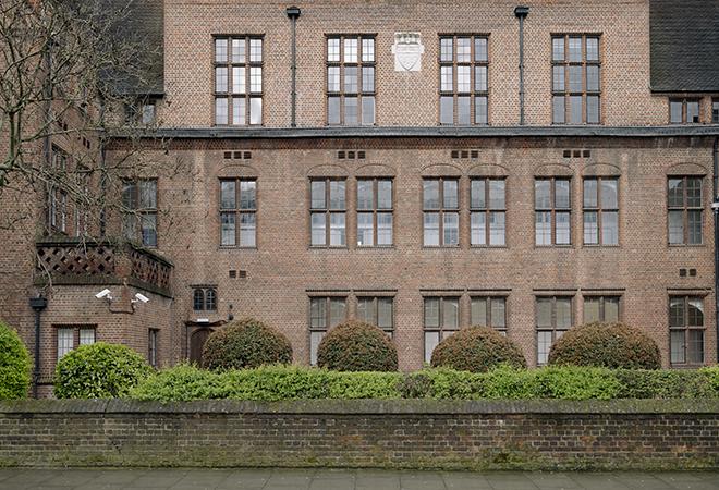 “Brick building with multiple rows of tall rectangular windows, a small shield emblem near the roof, and neatly trimmed round bushes in front of a low brick wall along the sidewalk.”