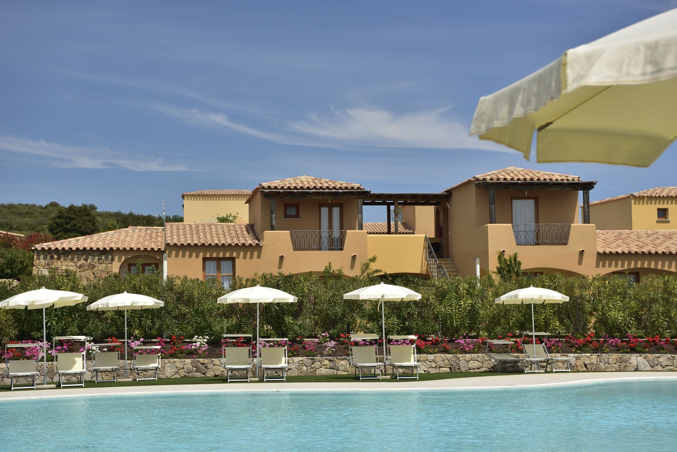 View from the pool area of a number of sun loungers and umbrellas with a small part of some hotel rooms behind on a clear sunny day.