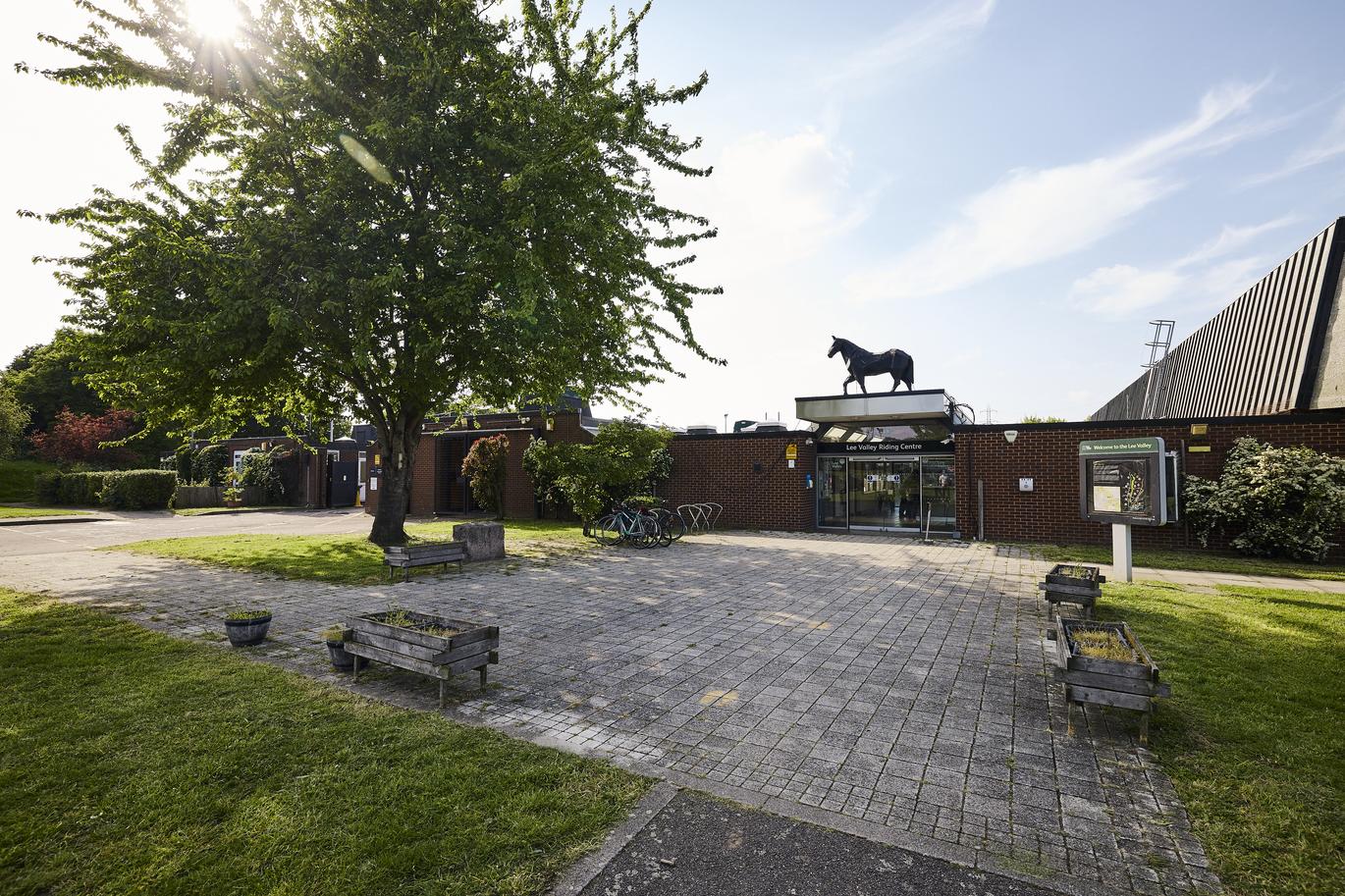 "Entrance to a riding centre with brick buildings, glass doors, and a horse sculpture above, surrounded by a paved courtyard and trees."
