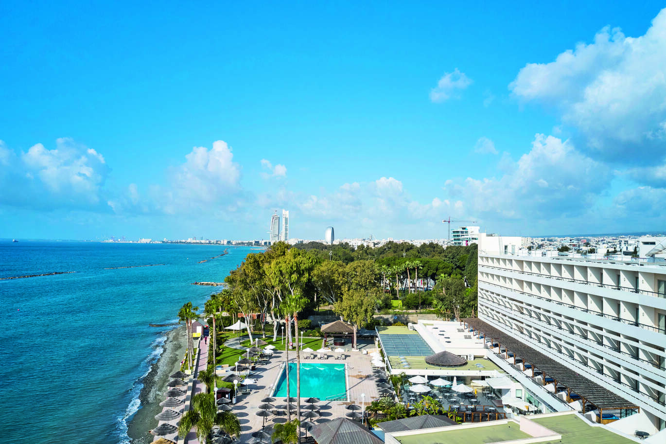 Aerial view of the hotel complex right on the beach, with the swimming pool in the middle with beds and umbrellas around and trees surrounding. With the hotel building behind the pool area and tall buildings in the background.