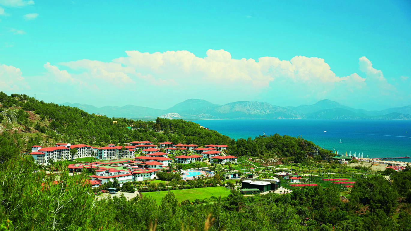 Long aerial shot of the resort surrounded by much greenery and trees with the blue sea behind with many boats in and mountains in the background the other side of the sea with some fluffy clouds in the blue sky above.