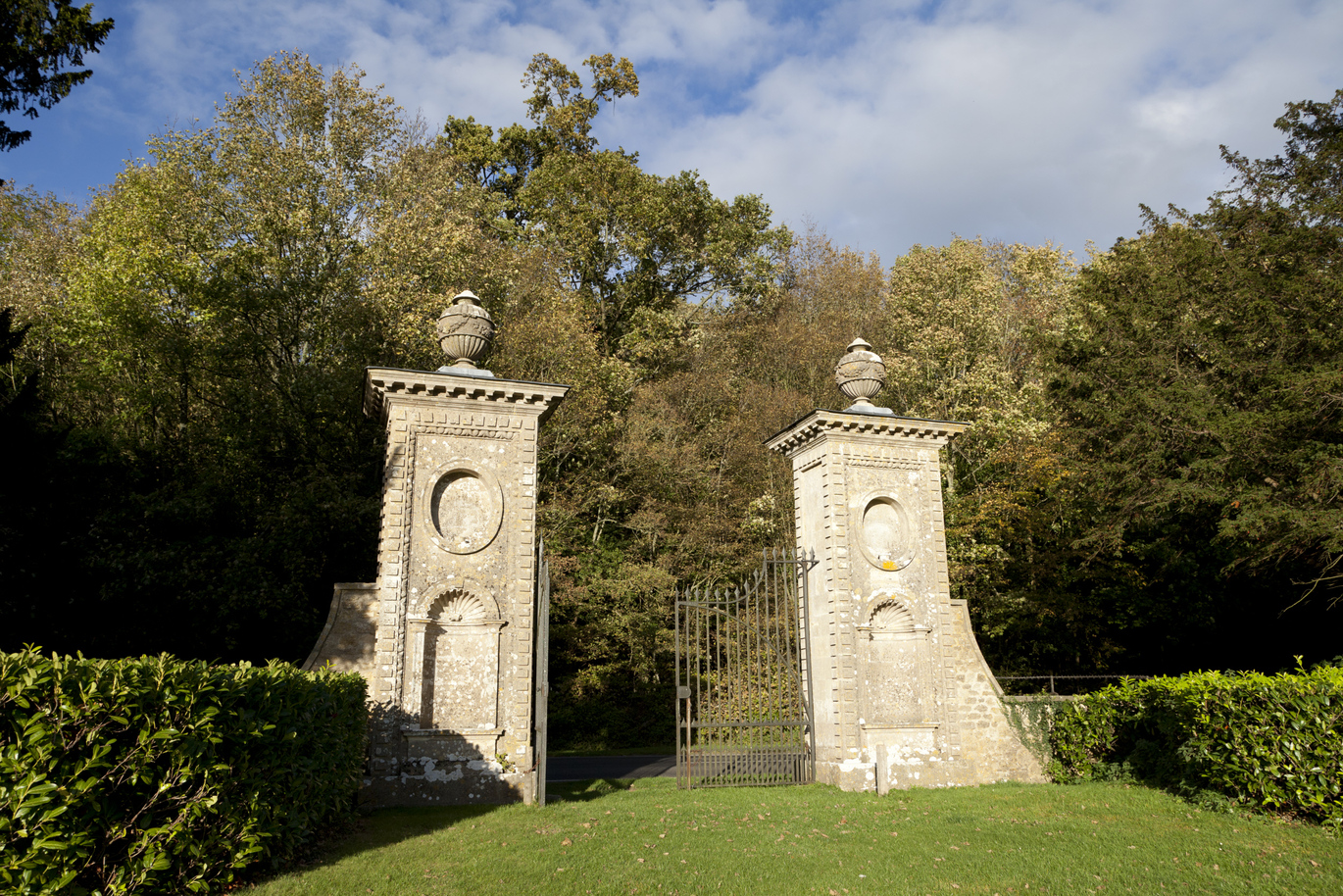 The large stone Gateposts of Coleshill Park with black iron gates, surrounded by many many trees and lush green lawn.