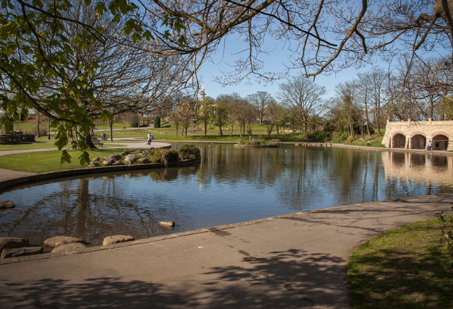 A picture of a lake with a bridge to the right of the image with arches underneath. There are some large stepping stone like rocks in the left hand corner of the lake. There is lots of green grass and trees all around, there is a path through the park with people walking along it. The sky is lovely and blue.