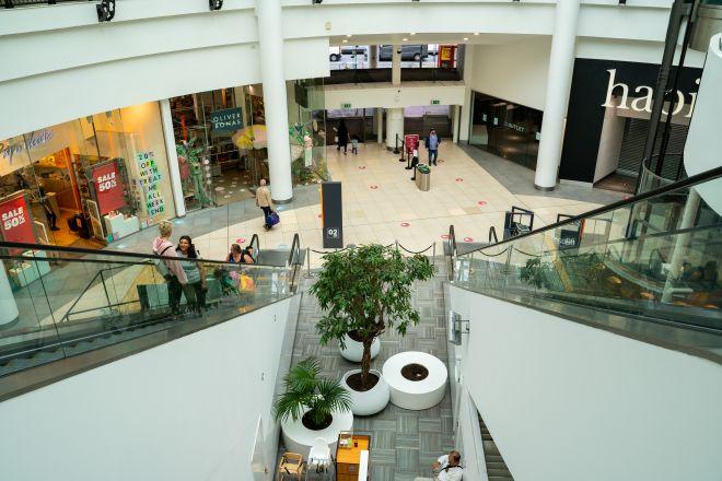 "Shopping centre atrium with escalators, upper-level walkway and central seating area with plants."