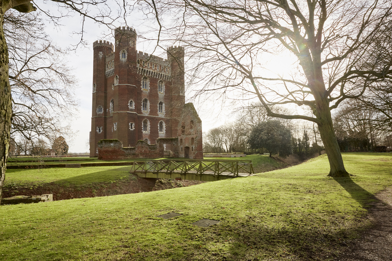 The west front of the 15th century Great Tower with the Stable ruins at Tattershall Castle. Surrounded by green lawns with a small wooden bridge over a ditch.