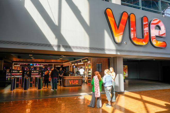 "Interior entrance to Vue cinema at Xscape with large illuminated sign, ticket kiosks and visitors standing nearby."