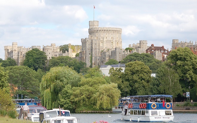 "Photo of river boats on the river, in the background is Windsor Castle with trees in front of it to the side of the river. The sky is overcast with many clouds"